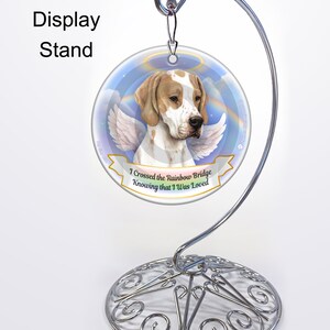 May include: A white and brown dog with angel wings on a round ornament with a blue sky and rainbow background. The ornament has the text "I crossed the Rainbow Bridge Knowing that I was Loved". The ornament is on a silver stand.