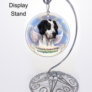 May include: A round ornament featuring a black and white dog with angel wings, displayed on a silver metal stand. The ornament has the text "I Crossed the Rainbow Bridge Knowing that I Was Loved". The stand has a decorative, scrolled base.