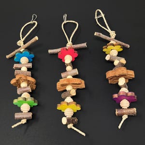 May include: Three colorful bird toys hanging against a black background. Each toy features wooden elements, including small branches, wooden beads, and flower-shaped accents in blue, red, yellow, green, and purple. The toys are suspended by natural twine.