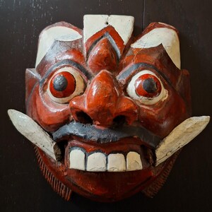 Op de afbeelding: Een gesneden houten masker, rood geschilderd met witte details. Het masker heeft een woeste uitdrukking, met grote, ronde ogen, een prominente neus en een zwarte snor. De mond staat open en toont witte tanden en grote witte slagtanden. Traditioneel ambachtelijk item.