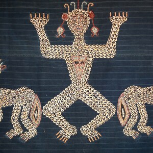 May include: A textile wall hanging with a central figure and two flanking figures, all made of shells. The figures are stylized and have a human-like form. The central figure has raised arms and a crown of shells. The background is a dark blue fabric.