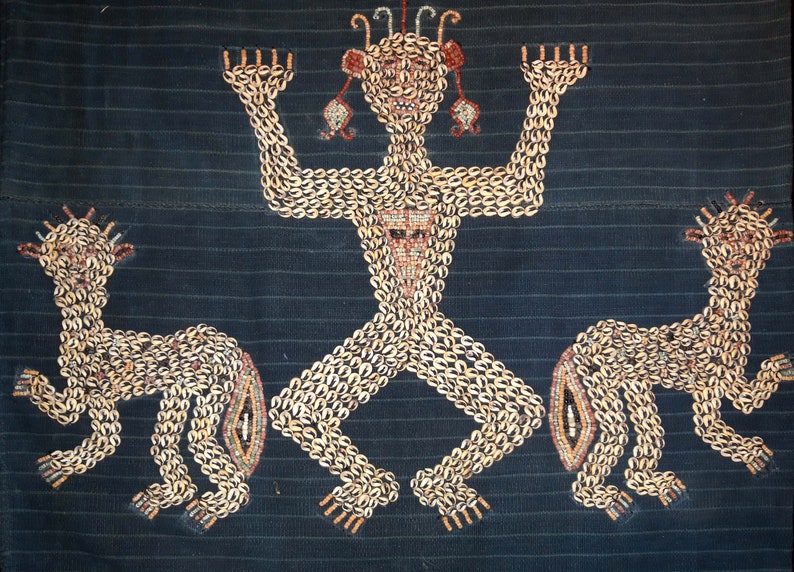 May include: A textile wall hanging with a central figure and two flanking figures, all made of shells. The figures are stylized and have a human-like form. The central figure has raised arms and a crown of shells. The background is a dark blue fabric.