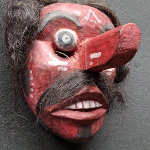 May include: A hand-carved wooden mask with a red face, large nose, and black mustache and hair. The mask has white teeth and a painted design above the eye. The mask is likely for theatrical or decorative purposes.