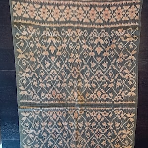 May include: A traditional woven textile with a geometric pattern in shades of brown, beige, and white. The fabric has a fringed edge.