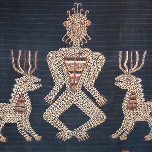 May include: A textile artwork depicting a human figure with two deer flanking it. The artwork is made of small, white shells sewn onto a dark blue fabric. The human figure has a crown of shells and a geometric pattern on its chest.