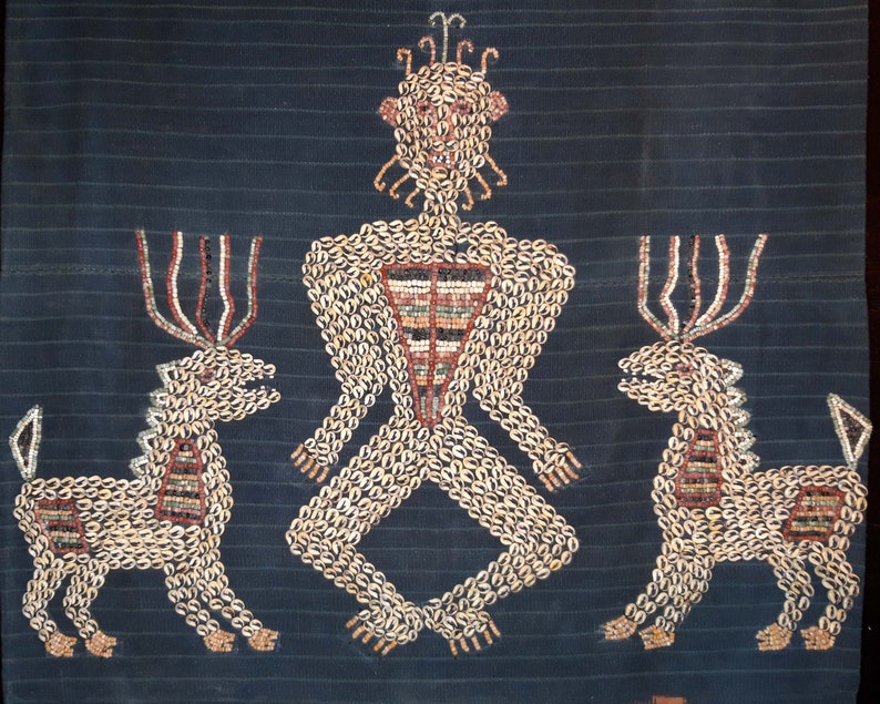 May include: A textile artwork depicting a human figure with two deer flanking it. The artwork is made of small, white shells sewn onto a dark blue fabric. The human figure has a crown of shells and a geometric pattern on its chest.