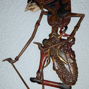Op de afbeelding: Een traditionele Indonesische schaduwpop, bekend als wayang kulit, die een personage voorstelt met een zwart en rood kostuum, dat een wapen vasthoudt. De pop is gemaakt van gesneden hout en beschilderd met ingewikkelde details. De pop is bevestigd aan een houten stok en is klaar om te worden gebruikt in een schaduwpoppenvoorstelling.