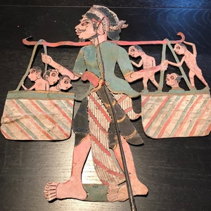 Op de afbeelding: Een handgeschilderde schaduwpop die een figuur voorstelt die twee manden draagt ​​gevuld met kleinere figuren. De hoofdfiguur draagt ​​een groen shirt en een rood-witte gepatroneerde broek. De manden zijn gestreept met rood en wit.