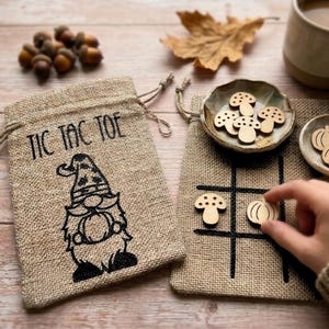 Könnte beinhalten: Ein Tic Tac Toe-Spielset aus Jute mit einem Kordelzugbeutel mit einem Zwerg-Design. Das Spiel enthält Spielsteine aus Holz in Form von Pilzen und Kürbissen. Eine Hand platziert ein Kürbisstück auf dem Spielbrett.