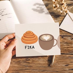 Pode incluir: Um cartão de felicitações retangular com uma ilustração gráfica de um rolo de canela dourado e uma caneca de café castanha com um design de espuma em forma de coração. A palavra FIKA está impressa em letras maiúsculas pretas sob o rolo.