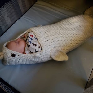 May include: A baby sleeping in a white crocheted narwhal-shaped swaddle blanket. The blanket has a white crocheted horn and a black button for an eye.