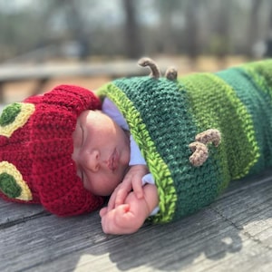 May include: A baby wearing a red and green crocheted caterpillar costume. The baby is sleeping and has a green and brown crocheted blanket wrapped around them.