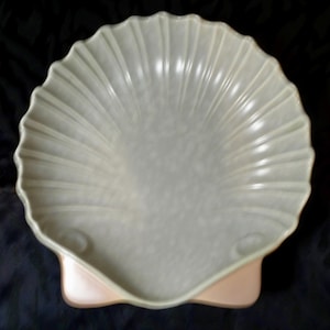 Puede incluir: Una fuente grande de cerámica gris claro con forma de concha de vieira. La fuente tiene un diseño acanalado y se apoya sobre una base rosa pálido. La fuente con forma de concha es una pieza decorativa.