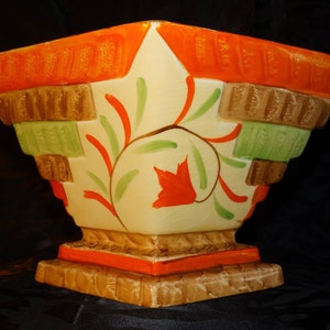 Könnte beinhalten: Ein antiker Keramik-Pflanztopf mit geometrischem Design in Orange-, Braun- und Grüntönen. Der Topf hat ein stilisiertes Blumenmotiv und einen quadratischen Sockel.