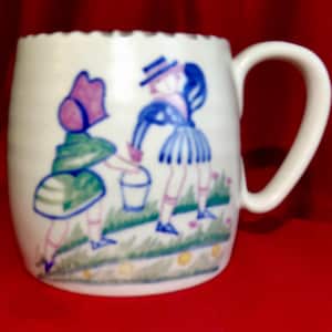 May include: A white ceramic mug featuring a hand-painted illustration of two figures in a garden setting. One figure wears a green dress and purple hat, while the other wears a blue striped outfit and hat. They are depicted near a bucket.