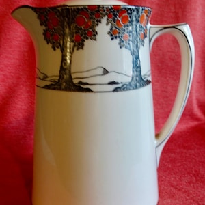 May include: A white ceramic jug with a lid, decorated with a design of trees bearing red fruit and blue leaves. The handle and rim are outlined with a thin blue line. A classic kitchenware item.