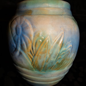 Può includere: Un vaso in ceramica con un corpo ampio e arrotondato e un bordo leggermente svasato. Il vaso presenta una superficie strutturata con un disegno di foglie stilizzate nelle tonalità del blu, verde e oro. La tavolozza dei colori è tenue, con una miscela di azzurro, crema e verde.