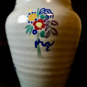 Puede incluir: Un jarrón de cerámica de color claro con un diseño floral pintado. Las flores son rojas, amarillas, azules y moradas, con hojas verdes y una cinta azul. El jarrón tiene una superficie texturizada y una forma ligeramente acampanada.