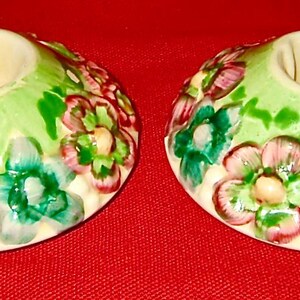 May include: A pair of round ceramic candle holders featuring a light green base decorated with raised, hand-painted floral motifs in shades of pink, teal, and white. Each holder has a central circular opening designed to accommodate a taper candle.