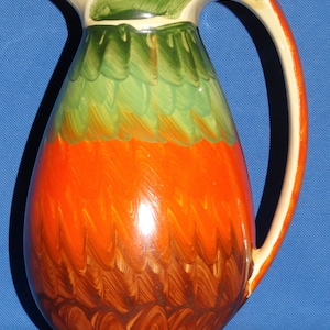 Puede incluir: Una jarra de cerámica con un diseño pintado en marrón, naranja, verde y blanco. La jarra tiene un cuerpo ancho y redondeado y un asa curva.