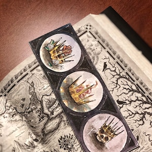 May include: A bookmark featuring three watercolour illustrations of crowns. The crowns are gold with red jewels and are surrounded by a white circle. The bookmark is on a map with the words "Sand Harbor", "Rolant", "The El", "Prynn", "The", "Stonegall", "Dow", "Capital City", "Bardon Harbor", "Bastiang", and "The Warrior's City" visible.