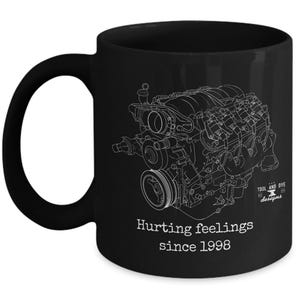 Puede incluir: Taza de cerámica negra con un dibujo lineal blanco de un motor. La taza tiene el texto "Hurting feelings since 1998" y el logotipo "Tool and Dye designs". La taza es de tamaño estándar y tiene un asa curvada.