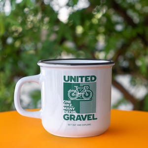 May include: White enamel mug with a black rim and handle. The mug features the text "UNITED GRAVEL" in green, along with a bicycle graphic. The mug also includes the text "ONE BIKE. MANY PATHS. GET OUT AND EXPLORE."