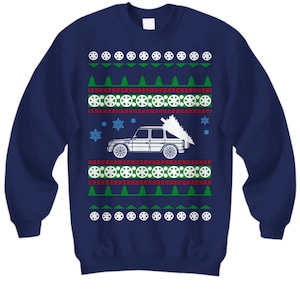 May include: A navy blue Christmas sweater with a white silhouette of a car carrying a Christmas tree. The sweater has a red and white snowflake pattern, green trees, and white tire designs.