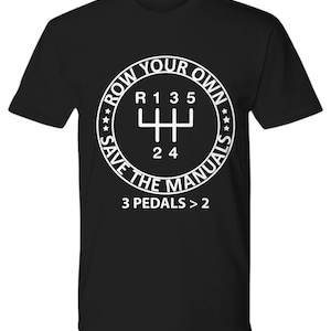 May include: Black t-shirt with a white circle graphic featuring a stick shift pattern and the text "Row Your Own Save The Manuals 3 Pedals > 2".