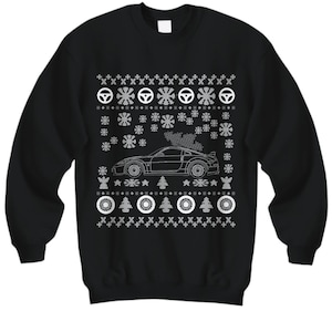 Könnte beinhalten: Schwarzer Pullover mit einem weißen Weihnachtsmotiv, das ein Auto mit einem Weihnachtsbaum auf dem Dach zeigt. Das Design enthält Schneeflocken, Sterne und Reifenspuren.