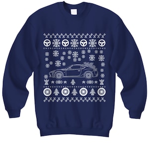 Könnte beinhalten: Ein marineblauer Pullover mit einem weißen Weihnachtsmotiv, das ein Auto mit einem Weihnachtsbaum auf dem Dach zeigt. Das Design enthält Schneeflocken, Sterne und Reifenspuren.