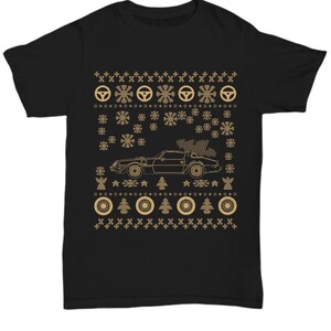 Puede incluir: Camiseta negra con un diseño dorado que presenta un coche con un árbol de Navidad, copos de nieve, volantes y otros elementos festivos. El diseño tiene un patrón similar al de un suéter navideño.