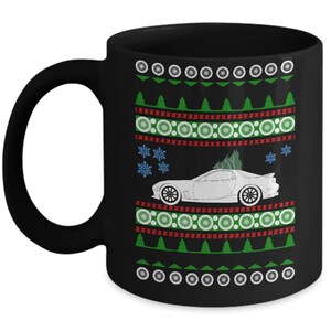 May include: Black ceramic mug with a festive design. The mug features a white car graphic with a Christmas tree on top, surrounded by green trees, snowflakes, and decorative patterns in red, green, and white. The mug has a black handle.