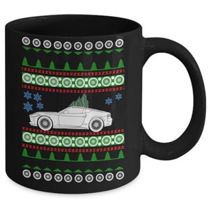 May include: Black ceramic mug with a festive design. The mug features a white car with a Christmas tree on top, surrounded by snowflakes, green trees, and red and green geometric patterns. The mug has a large, curved handle.
