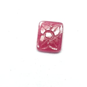 Può includere: Una piccola gemma rossa rettangolare con un motivo floreale intagliato. La gemma presenta un cerchio centrale circondato da quattro petali stilizzati. L'intaglio è dettagliato e la pietra ha una finitura lucida.
