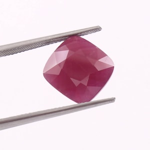 Może przedstawiać: Fasetowany rubin o szlifie poduszkowym trzymany pęsetą. Rubin ma głęboki, nasycony czerwony kolor z wieloma fasetami, które odbijają światło. Pęseta jest srebrna i trzyma kamień szlachetny na białym tle.