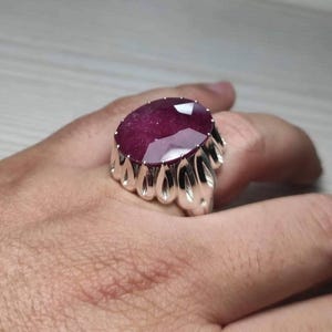 May include: A large, oval-cut ruby gemstone ring. The ruby is a deep red color and set in a silver-colored metal band with a unique, ornate design. The ring is shown on a hand.