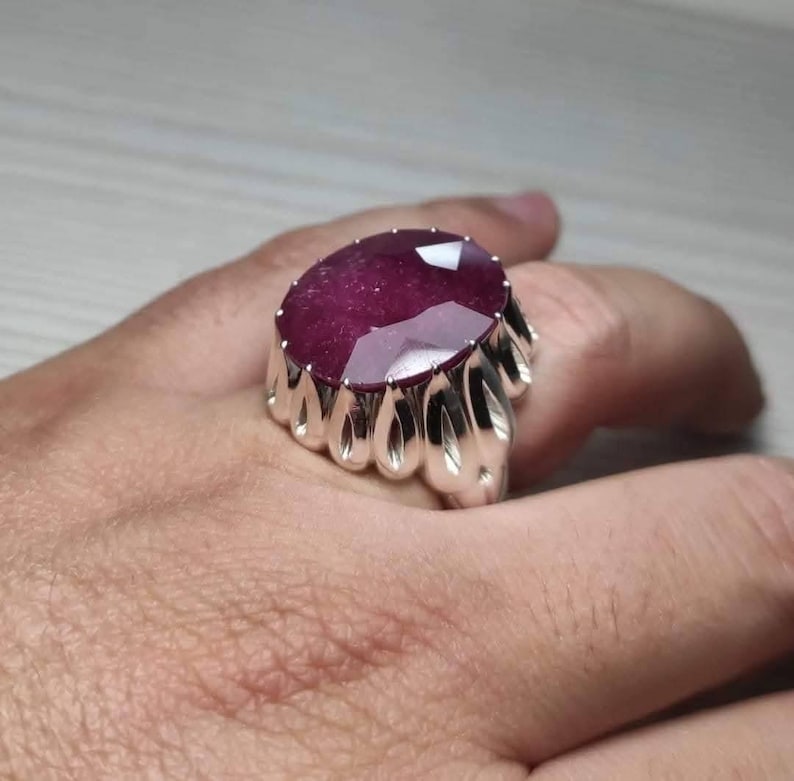 May include: A large, oval-cut ruby gemstone ring. The ruby is a deep red color and set in a silver-colored metal band with a unique, ornate design. The ring is shown on a hand.