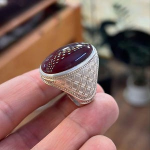 Puede incluir: Un anillo de plata con una piedra grande, ovalada y de color rojo oscuro. La banda del anillo presenta un patrón geométrico detallado. La piedra tiene una superficie pulida con un diseño sutil de puntos. El anillo se sostiene en una mano, mostrando su tamaño y diseño.