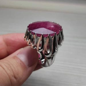 May include: A silver ring featuring a large, faceted, oval-shaped ruby gemstone. The ring has a decorative, openwork design around the ruby, with a polished finish. The ruby is a deep, rich red color.