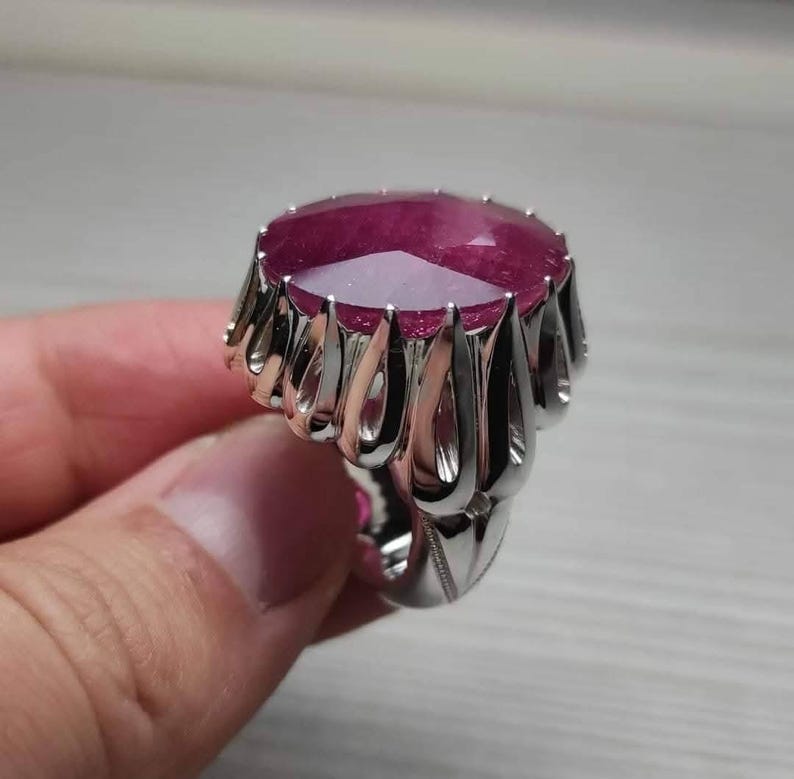 May include: A silver ring featuring a large, faceted, oval-shaped ruby gemstone. The ring has a decorative, openwork design around the ruby, with a polished finish. The ruby is a deep, rich red color.