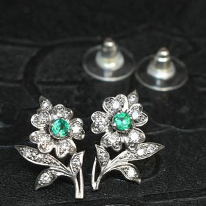 May include: A pair of flower-shaped stud earrings. Each earring features a central emerald-green gemstone surrounded by small, clear gemstones. The earrings are made of silver-colored metal and have a floral design with leaves.