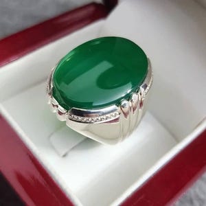 Puede incluir: Anillo de plata con una gran piedra verde ovalada pulida. El anillo tiene una banda de plata detallada con un diseño texturizado, presentado en una caja roja y blanca.