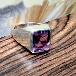 May include: A silver ring with a large, rectangular, deep purple amethyst gemstone. The ring has a simple, modern design with a thick band. The gemstone is the focal point, catching the light and showcasing its rich color. The ring is set on a wooden surface.