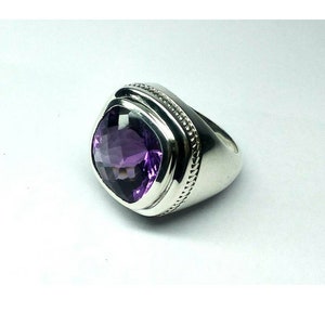 May include: A silver ring with a large, square-cut, deep purple amethyst gemstone. The ring has a thick band with a textured design around the gemstone setting. The ring is set against a white background.