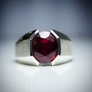 May include: A silver ring with a large, oval-cut ruby gemstone. The ruby is a deep red colour and is set in a polished silver band with a brushed texture on the sides. The ring has a modern, minimalist design.