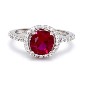 May include: A silver ring featuring a large, oval-cut, deep red gemstone, surrounded by a halo of smaller, clear stones. The ring's band is also adorned with small, clear stones, creating a sparkling effect. The ring is set against a white background.