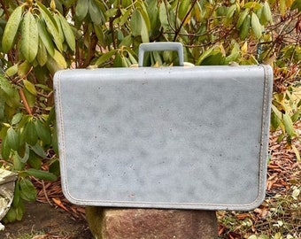 May include: A vintage gray suitcase with a handle. The suitcase is sitting on a rock.