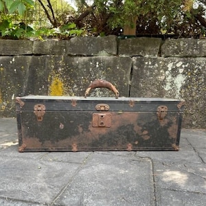 May include: An antique black trunk with a brown leather handle and rusty metal hardware. The trunk is rectangular and shows signs of age and wear, with visible rust and discoloration. The trunk is sitting on a gray stone surface.