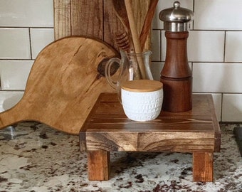 May include: A wooden tiered stand with a white ceramic jar with a wooden lid, a wooden pepper mill, and a glass pitcher on top. The stand is on a white tiled countertop.